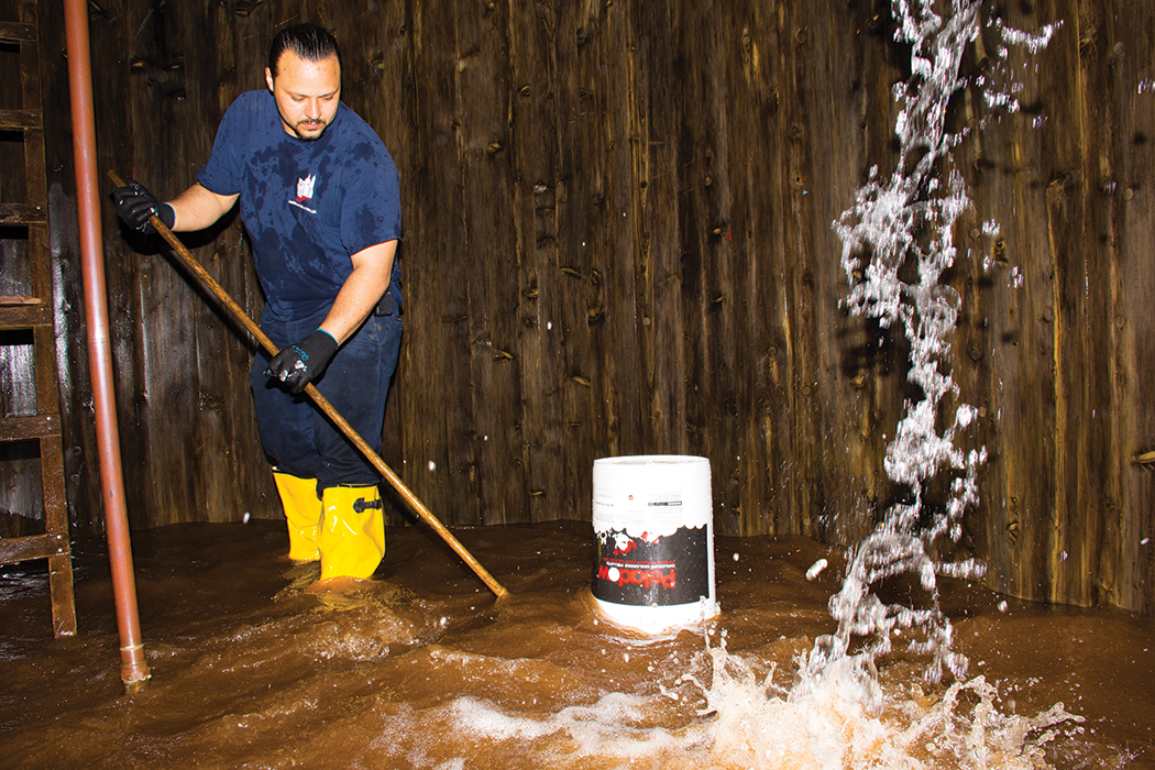 Water at the bottom of the tank is dark brown as Jonathan Lewin pushes the sediment toward the drain. An overturned bucket on top of the drain pipe makes a slurping sound as suction helps pull the muddy water out of the tank.